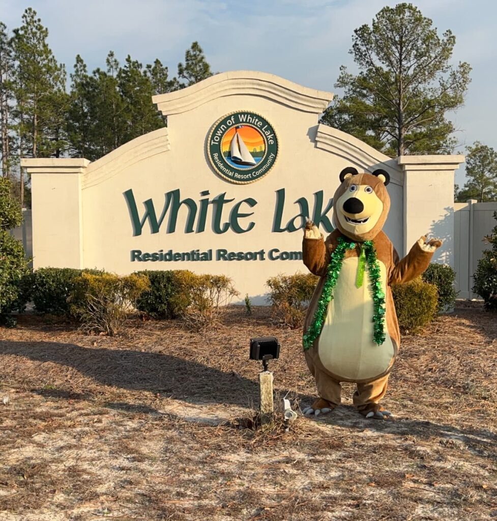 A cheerful bear mascot poses in front of the White Lake Residential Resort Community sign, surrounded by greenery.