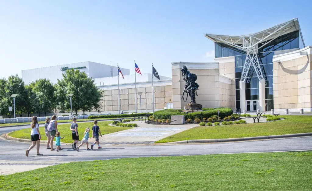A family walks towards a museum featuring a soldier statue and flags, surrounded by greenery on a sunny day.