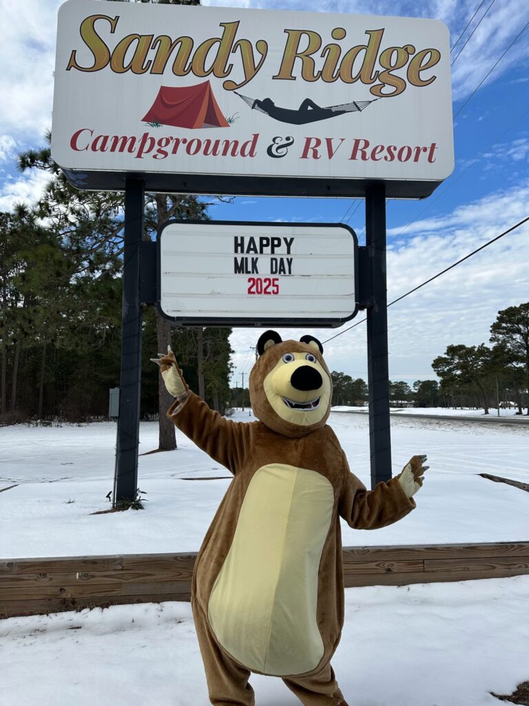 A cheerful bear mascot celebrating MLK Day 2025 at Sandy Ridge Campground and RV Resort under a blue sky.