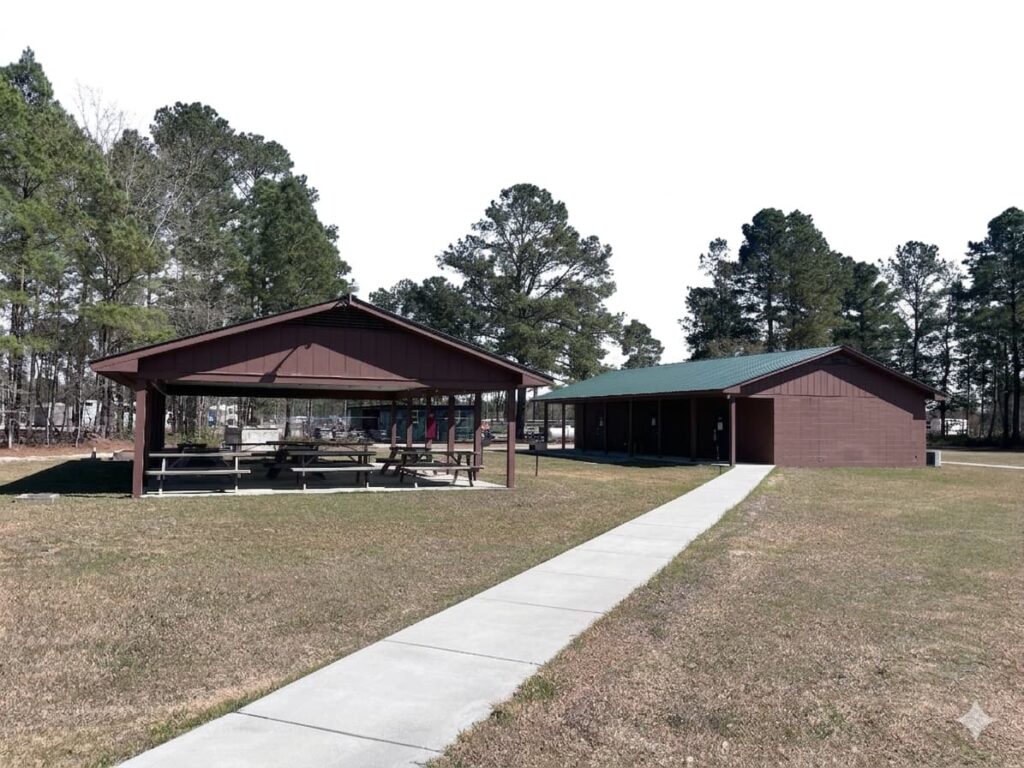 A spacious outdoor pavilion with picnic tables surrounded by trees, leading to a building in a park setting.