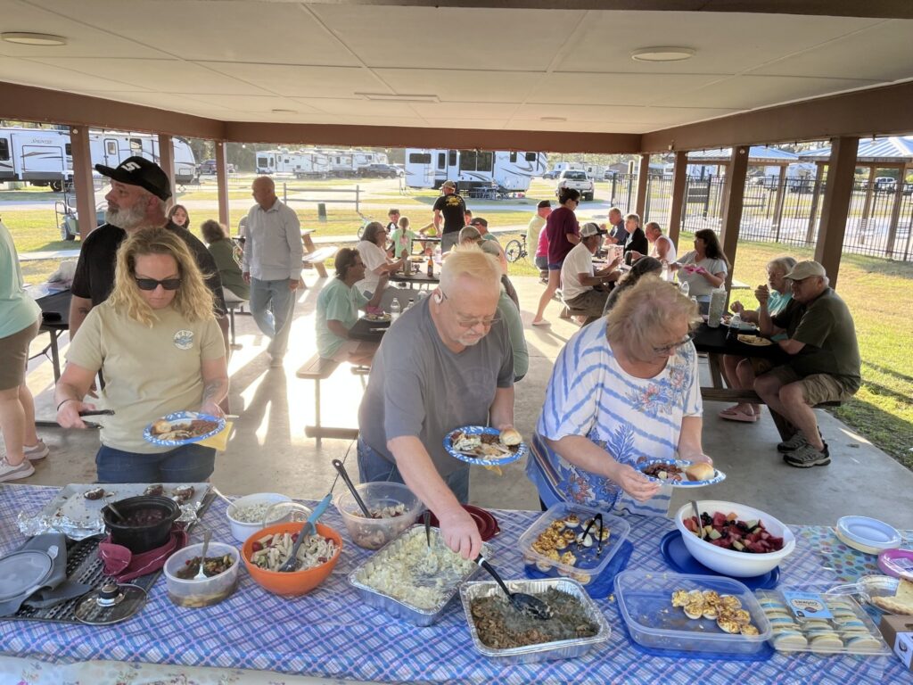 A community gathering with people enjoying a potluck meal under a pavilion, featuring various dishes and cheerful interactions.