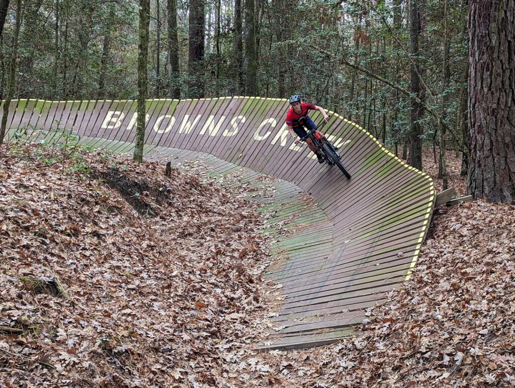 A cyclist skillfully navigates a wooden bike trail in a forest, surrounded by fallen leaves and trees.