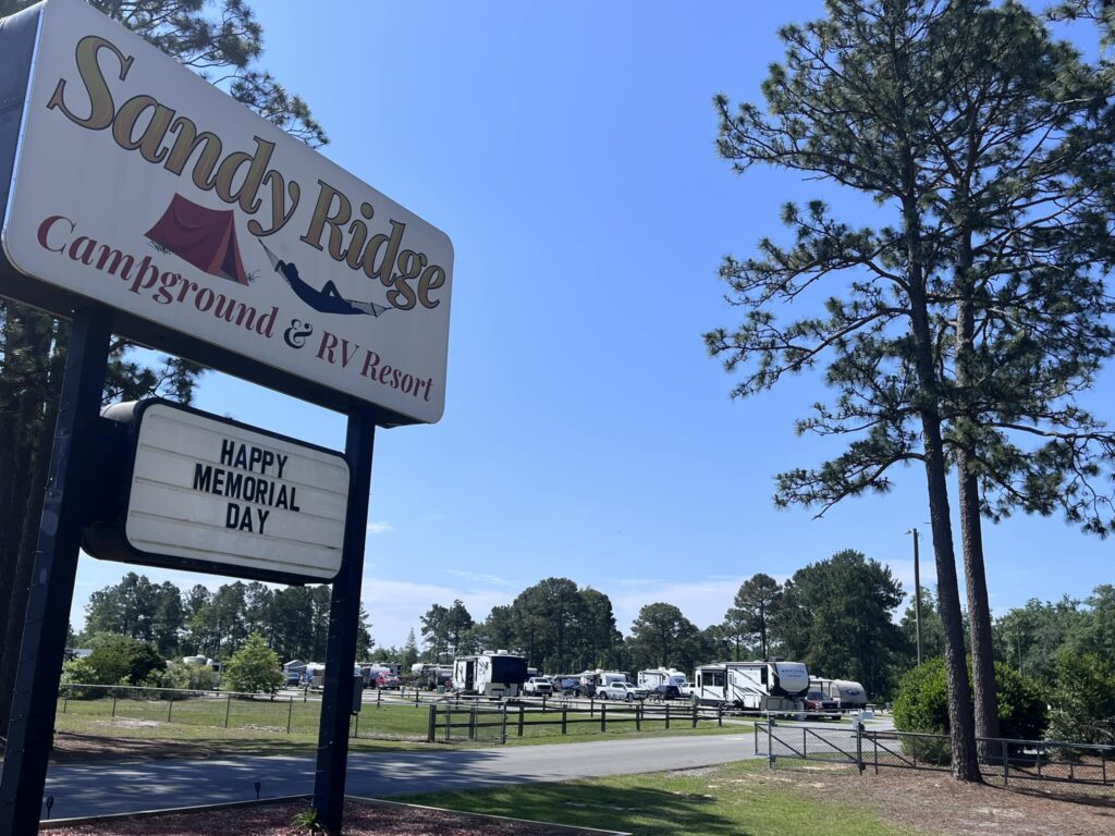 Sandy Ridge Campground and RV Resort sign with blue sky, trees, and RVs in the background, celebrating Memorial Day.