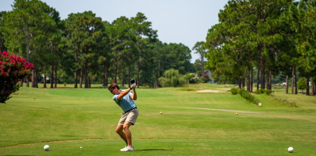 A golfer in a blue shirt swings a club on a lush green golf course surrounded by trees and flowers.