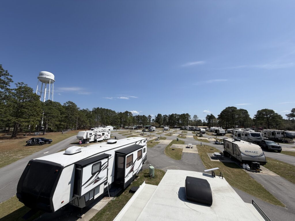 Aerial view of an RV park with multiple parked RVs, surrounded by trees and a water tower under a clear blue sky.