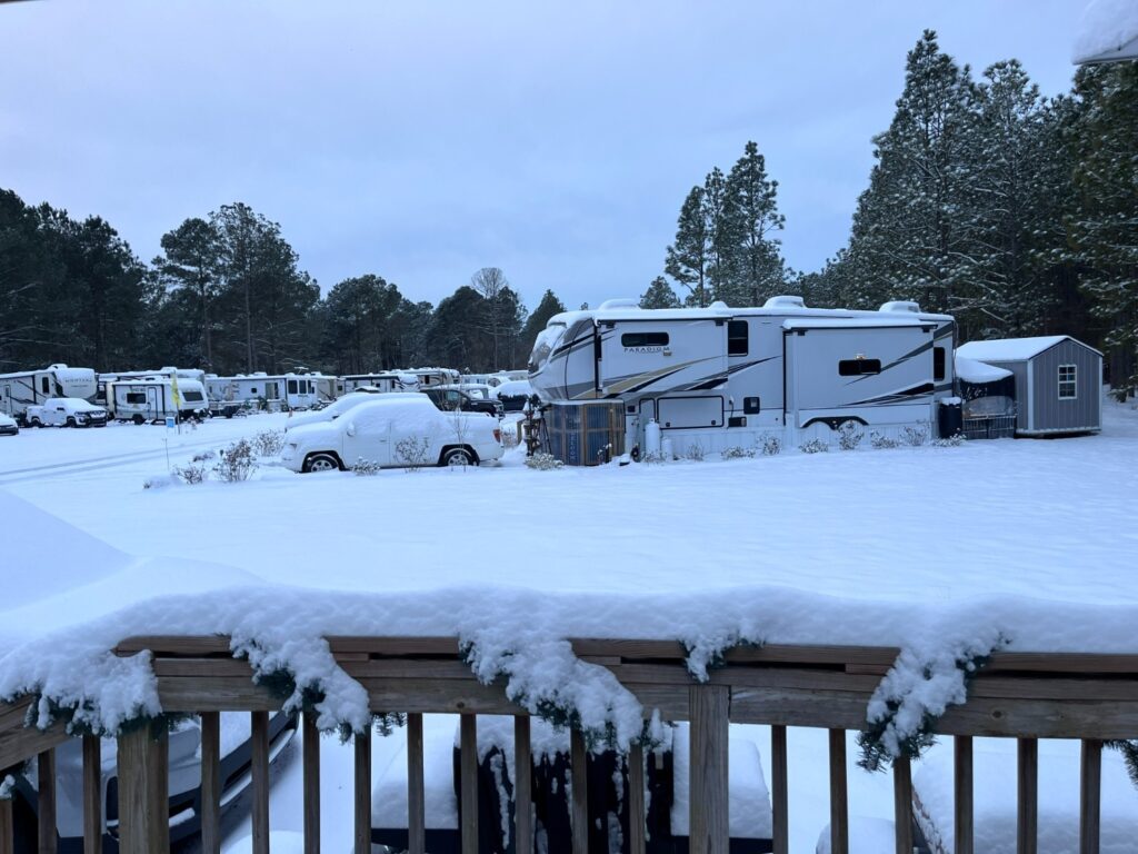 A snowy RV park scene featuring parked RVs and vehicles covered in snow, surrounded by trees under a cloudy sky.