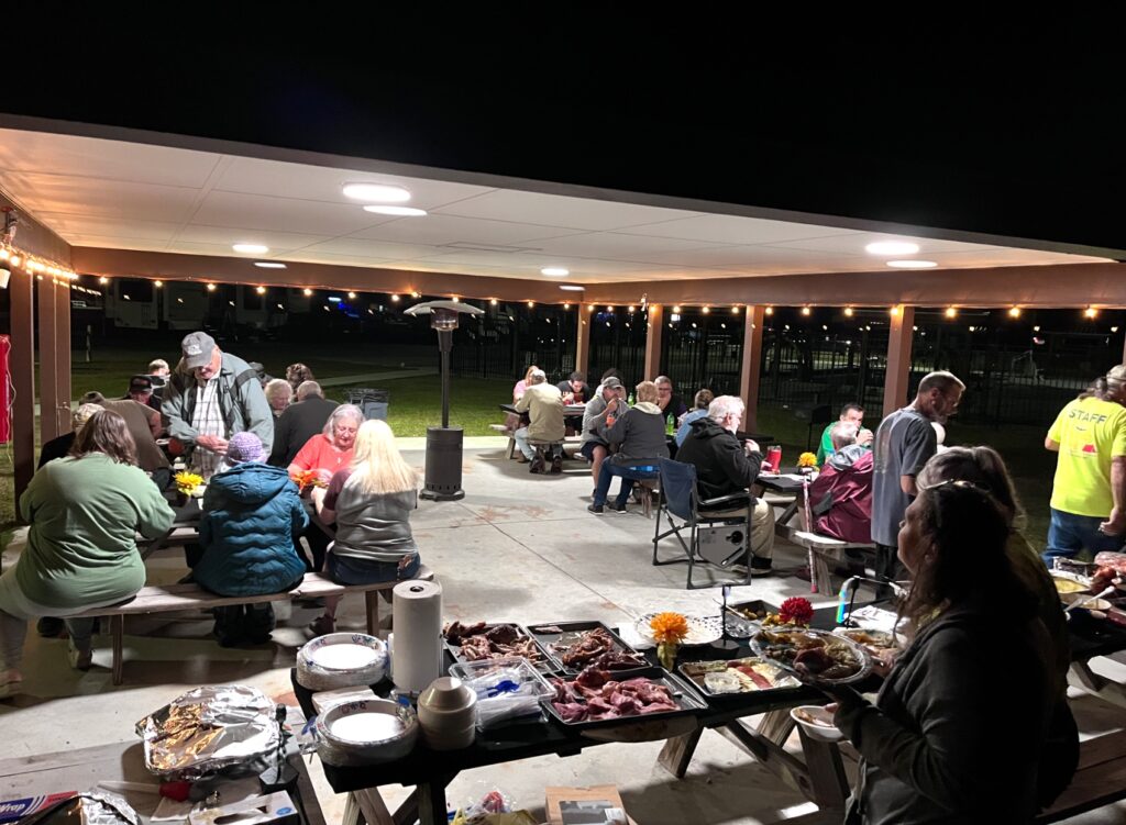 A lively outdoor gathering at night with people enjoying food and conversation under warm lights in a cozy setting.