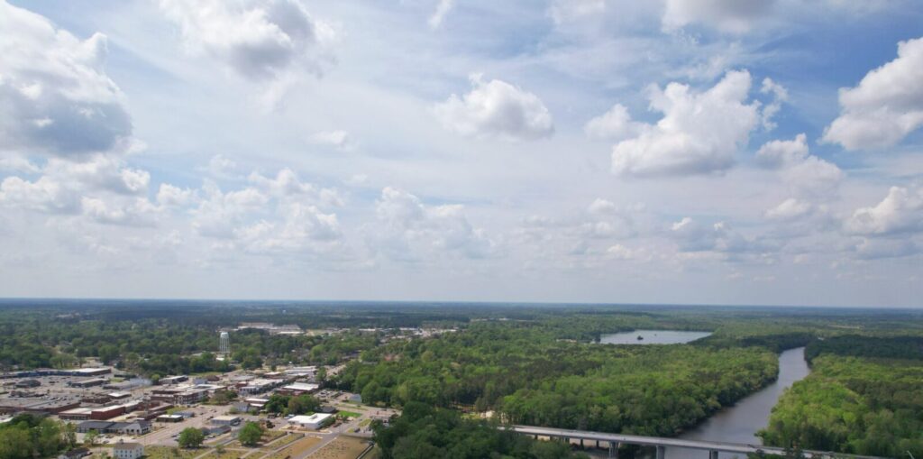 Aerial view of a lush green landscape with a river, clouds, and a small town in the distance under a bright sky.