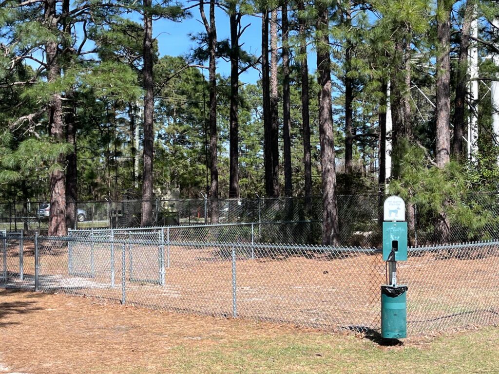 A fenced dog park surrounded by tall pine trees, featuring a waste disposal station and gravel ground.