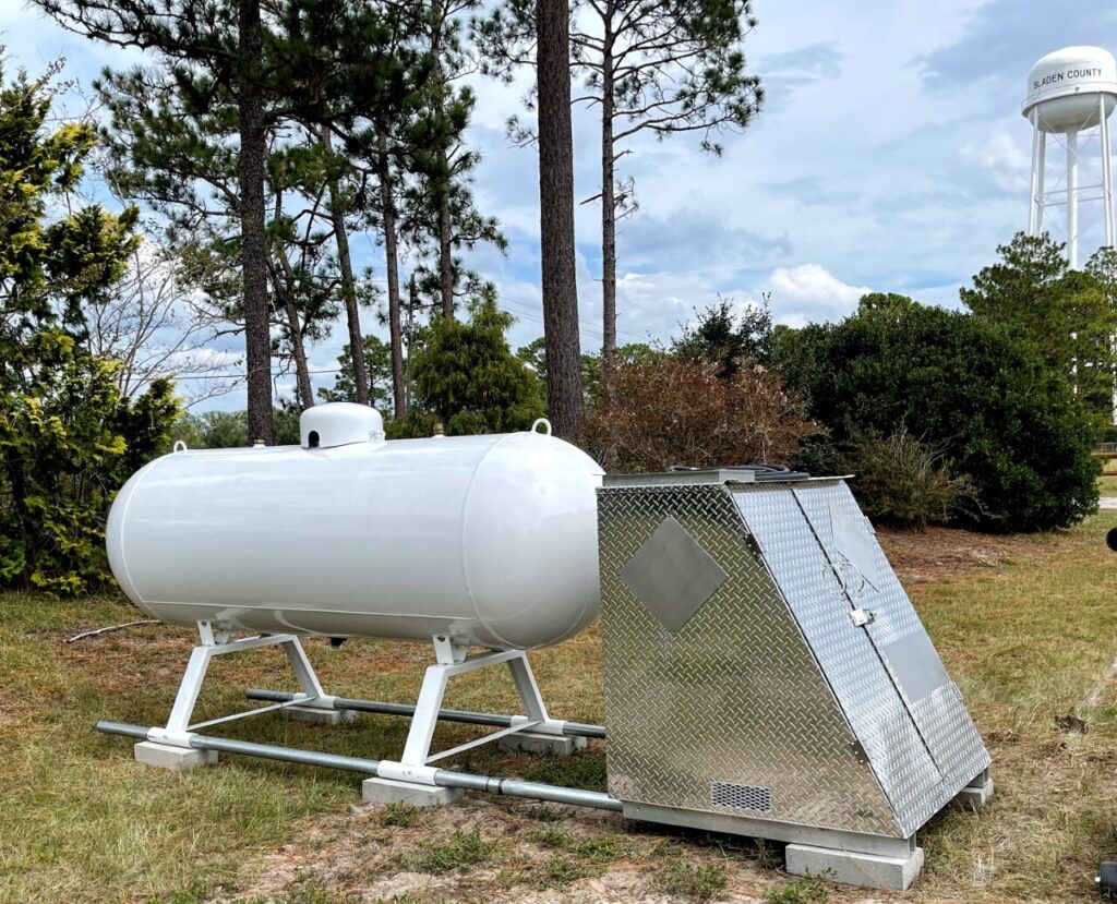 A white propane tank with a silver protective enclosure, surrounded by trees and a water tower in the background.
